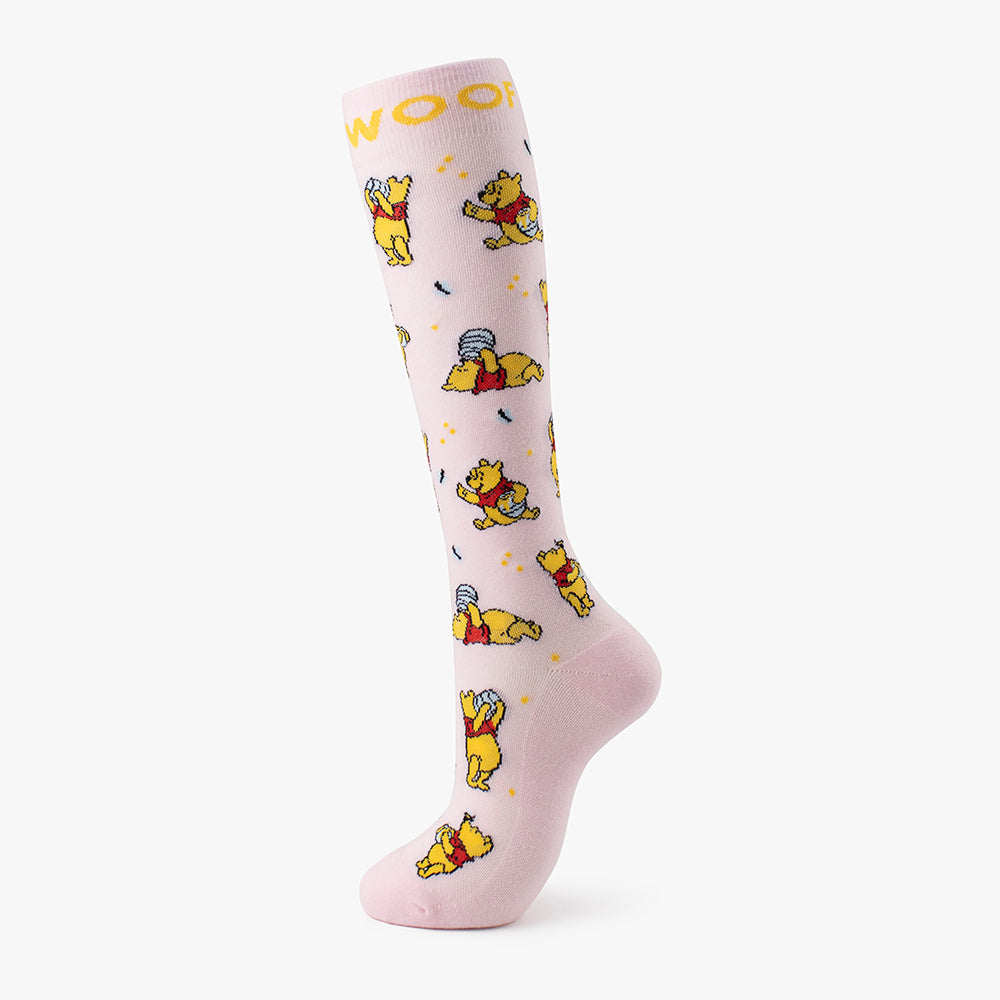 Studio shot of the soft pink pair from the Disney’s Winnie the Pooh Bamboo Compression Socks 4 Pack, demonstrating a gold "WOOF" cuff band, scattered hunny pots, and playful illustrations of Pooh eating honey.   