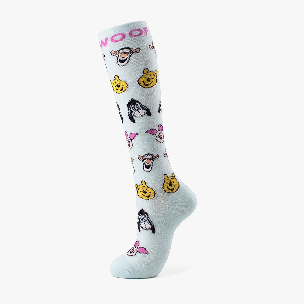 Studio shot of the light blue pair from the Disney’s Winnie the Pooh Bamboo Compression Socks 4 Pack, featuring a bright pink "WOOF" cuff band and an all-over repeating face motif of Pooh, Tigger, Eeyore, and Piglet.   